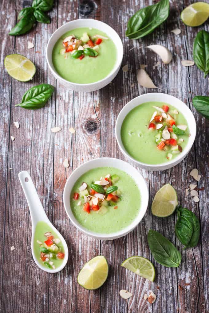 Three bowls of cold cucumber soup on a wooden table. Fresh basil leaves and lime wedges scattered around the bowls.