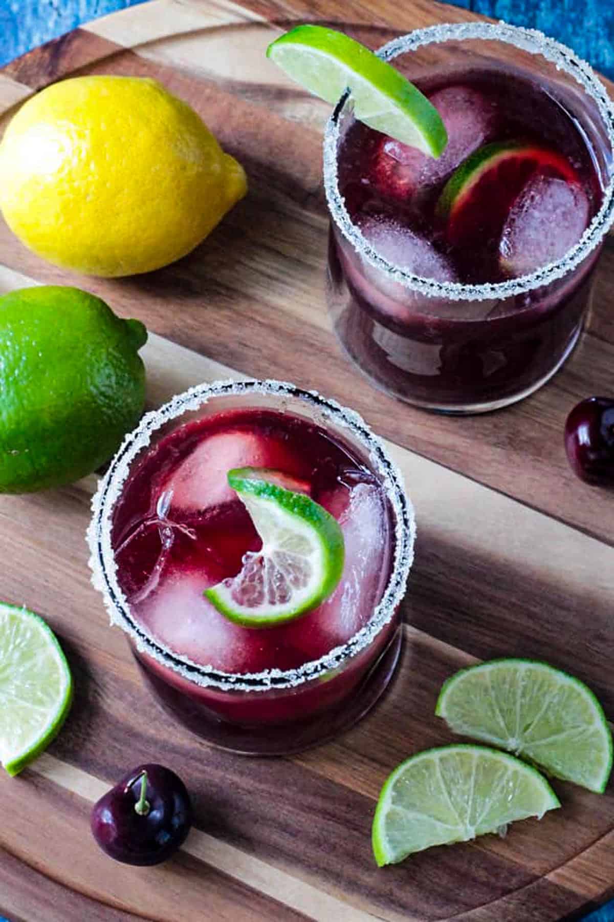 Two glasses of red juice drink on a circular wooden serving tray with lemons and limes.