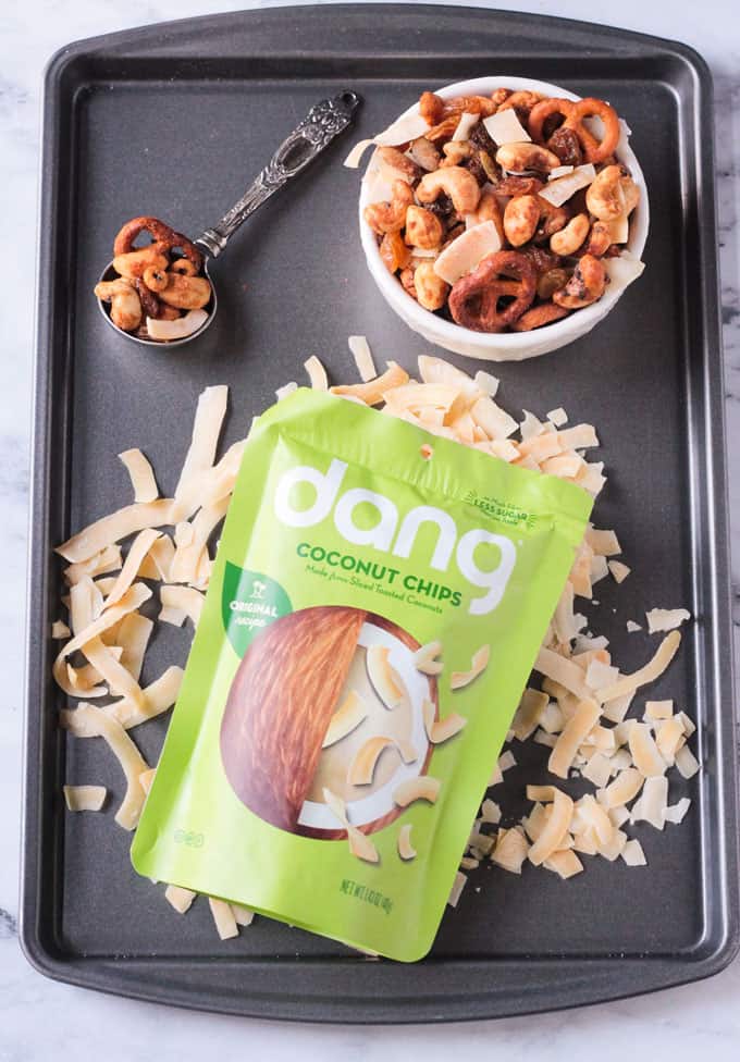 Overhead view of a bag of Dang coconut flakes lying on a bed of toasted coconut flakes on a baking sheet. Small bowl of trail mix behind also on the tray.