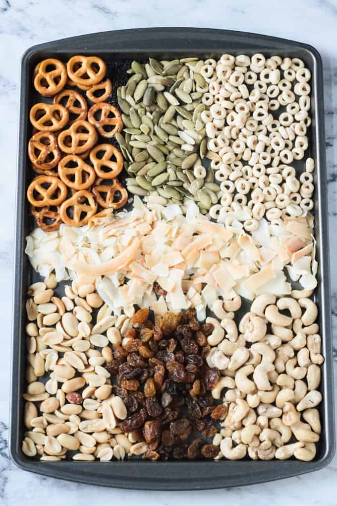 Baking tray with trail mix ingredients lined up - pretzels, pumpkin seeds, cheerios, coconut flakes, peanuts, golden raisins, and cashews.