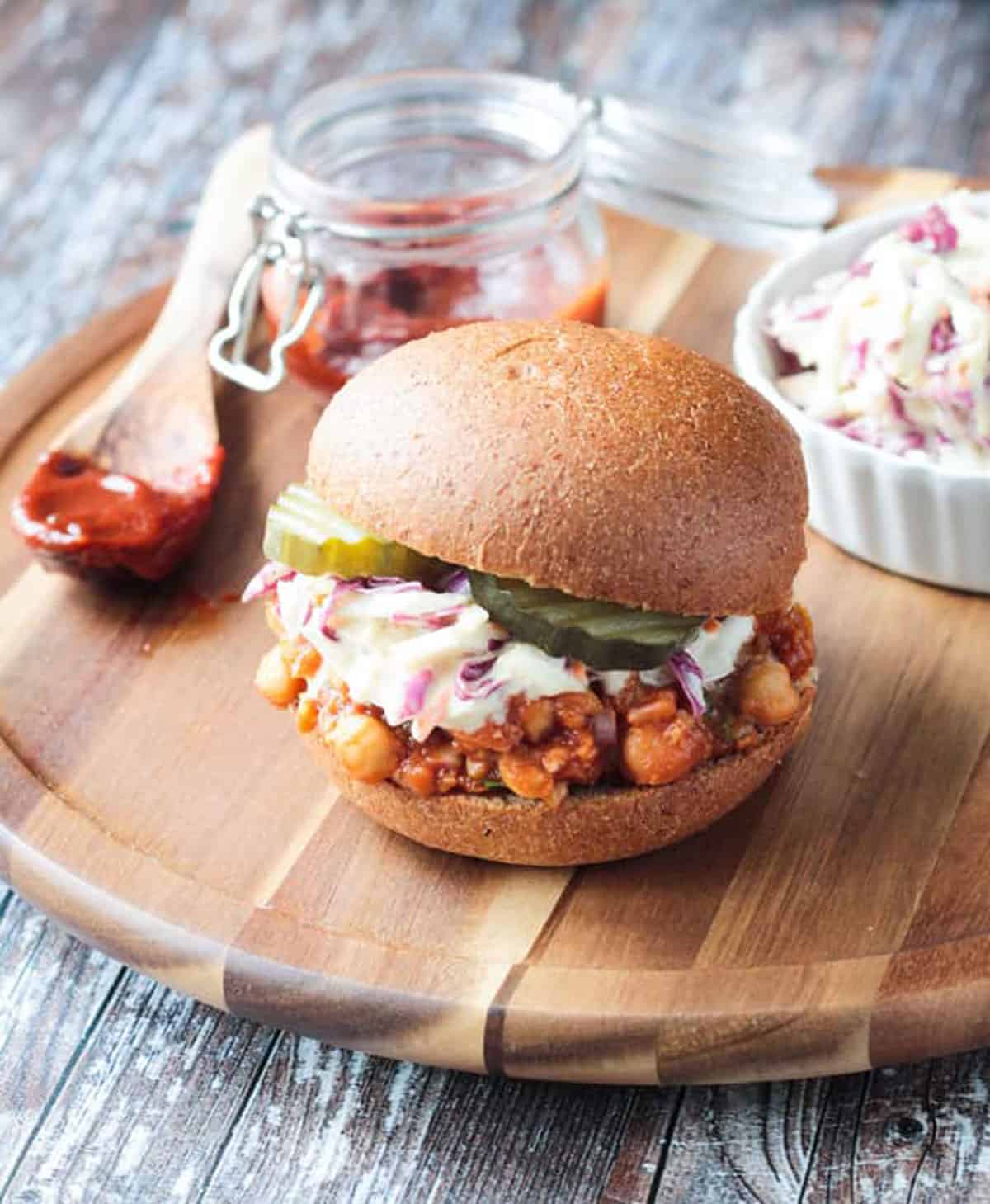 Vegan BBQ Sandwich on a round wooden platter with small jar of bbq sauce behind.
