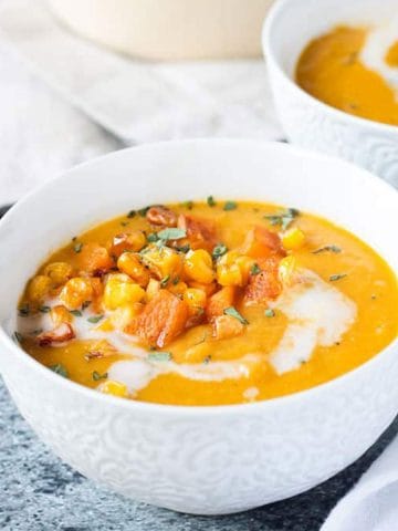 Close up of squash soup in a white bowl topped with diced squash, corn & thyme.