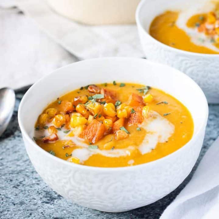 Close up of squash soup in a white bowl topped with diced squash, corn & thyme.