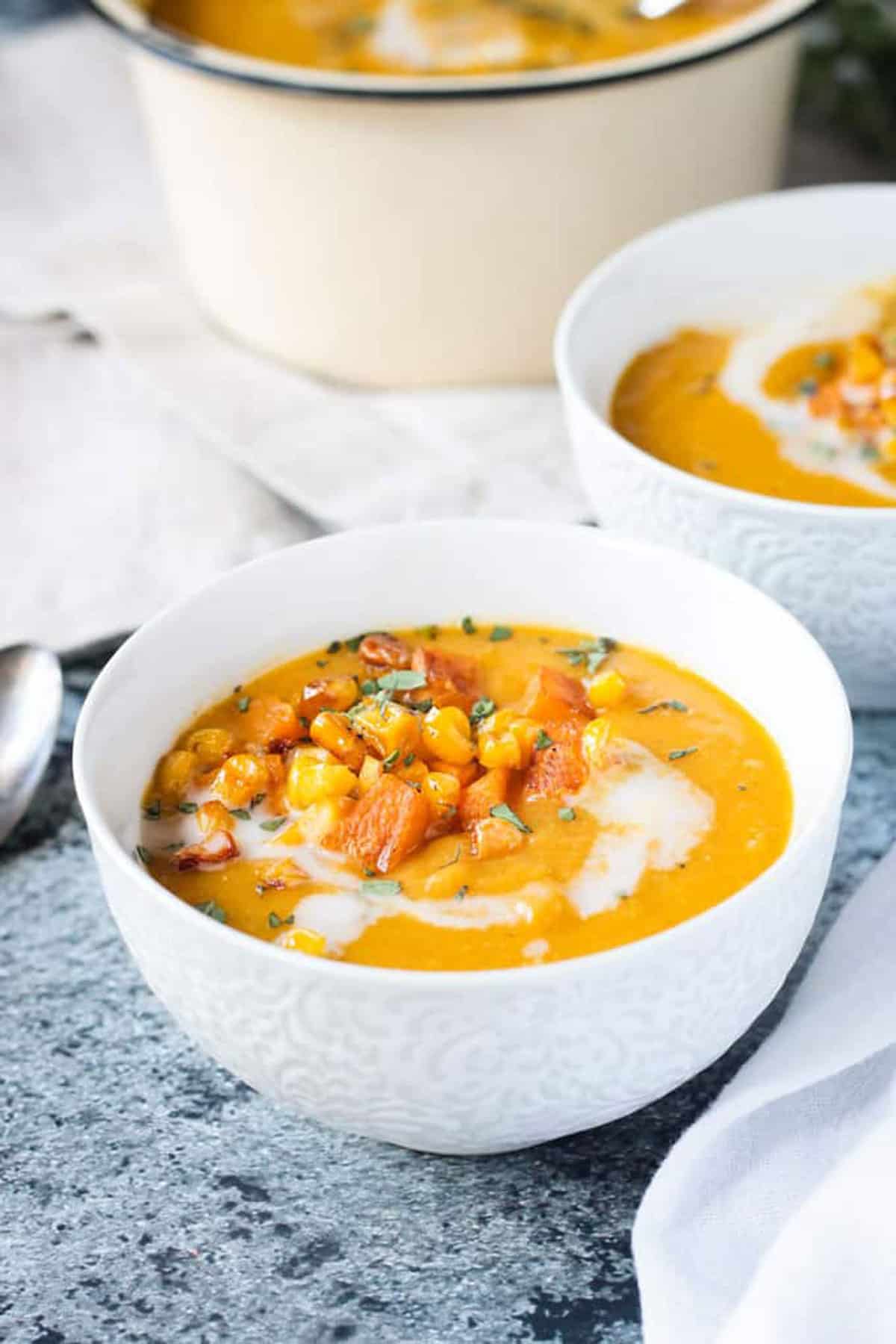 Close up of squash soup in a white bowl topped with diced squash, corn & thyme.