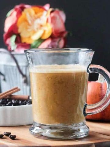 Front view of a glass of Pumpkin Latte. Orange decorative pumpkin and a small metal bucket of flowers in the background.