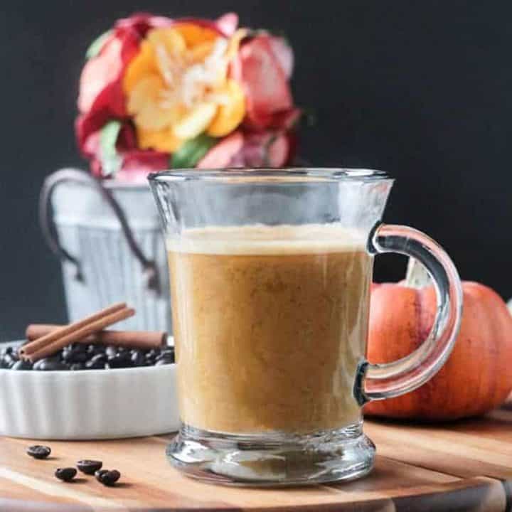 Front view of a glass of Pumpkin Latte. Orange decorative pumpkin and a small metal bucket of flowers in the background.