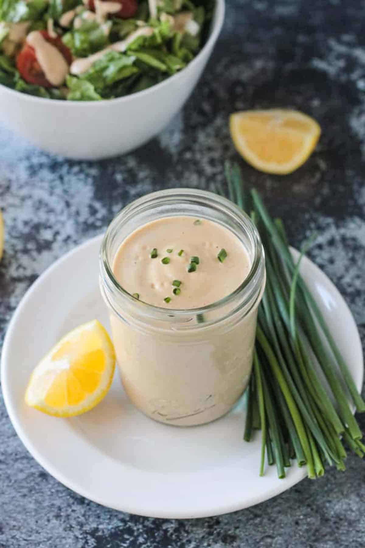 Vegan ranch dressing in a glass jar on a white plate next to chives and a lemon wedge.