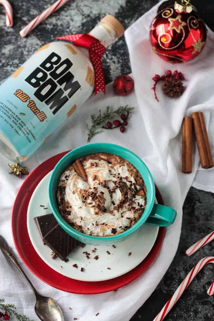 Overhead view of mug of spiked hot cocoa with a bottle of Bom Bom liquor next to it. Cinnamon sticks and candy canes lie nearby.