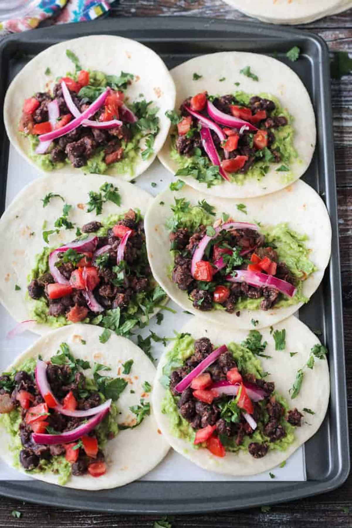 6 tortillas laid out flat with black beans, guacamole, pickled red onions, and cilantro.