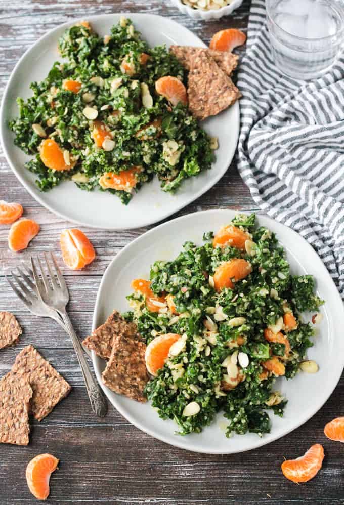 Two plates of massaged kale and quinoa salad with orange segments and forks lying near the plates.