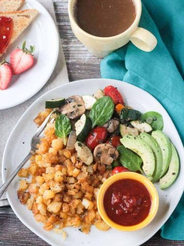 Metal fork on a plate with crispy breakfast hash browns, skillet vegetables, sliced avocado, and a small bowl of salsa.