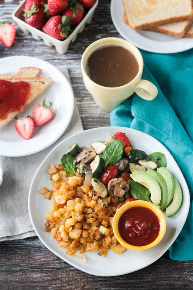 Overhead view of a vegan breakfast spread - plate of hash browns and veggies with a plate of toast and jam behind next to a cup of coffee.