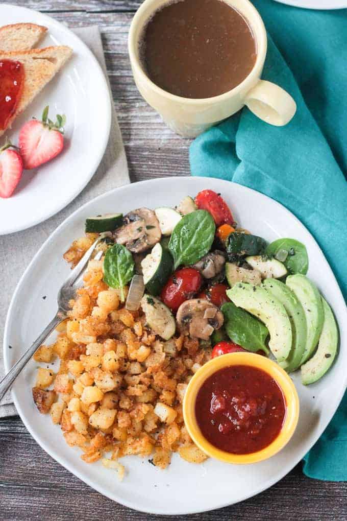 Metal fork on a plate with crispy breakfast hash browns, skillet vegetables, sliced avocado, and a small bowl of salsa.