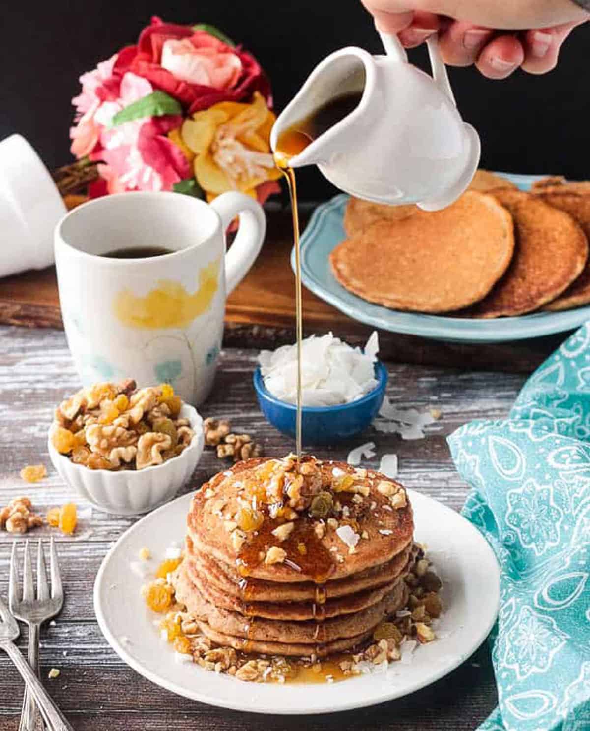 Syrup being poured onto a stack of pancakes from a small white pitcher.