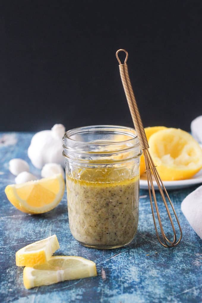 Lemon Vinaigrette Dressing in a glass jar with a gold wire whisk leaning up agains the jar and lemons in the background