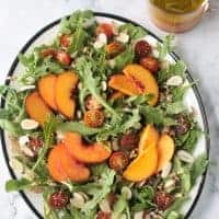 Platter of arugula salad topped with sliced peaches, halved grape tomatoes, slivered almonds, farro, and orange basil vinaigrette