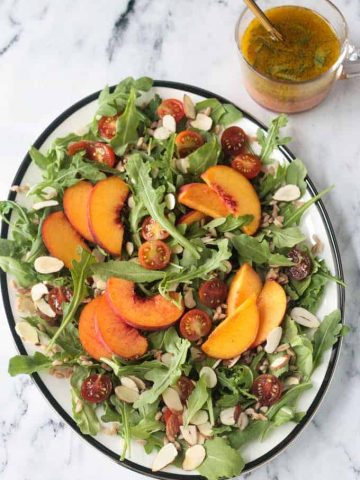 Platter of arugula salad topped with sliced peaches, halved grape tomatoes, slivered almonds, farro, and orange basil vinaigrette