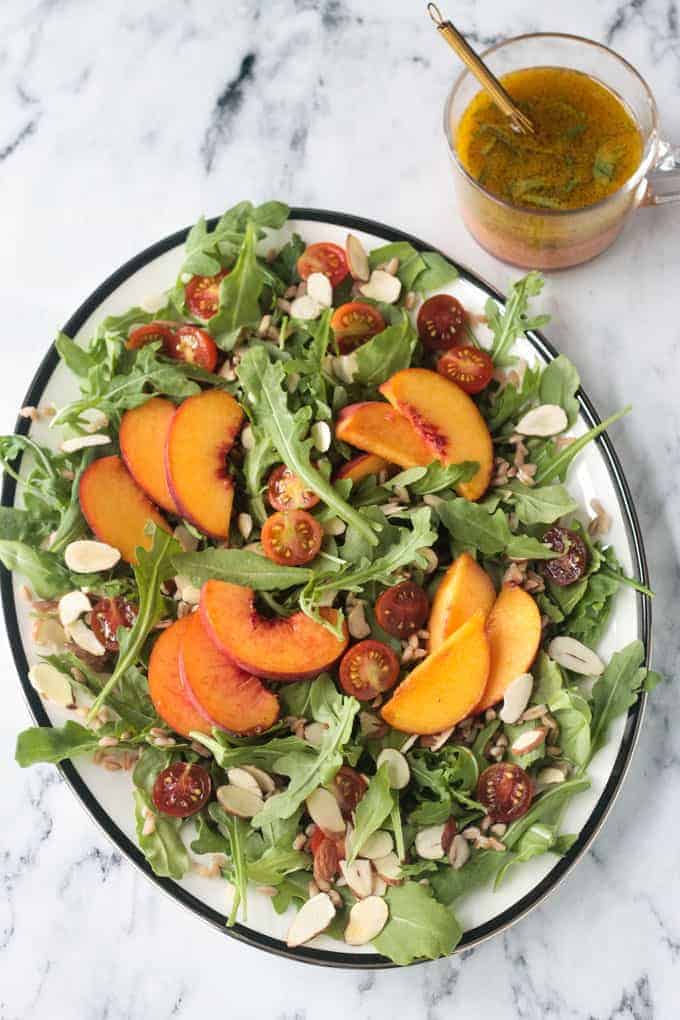 Platter of arugula salad topped with sliced peaches, halved grape tomatoes, slivered almonds, farro, and orange basil vinaigrette