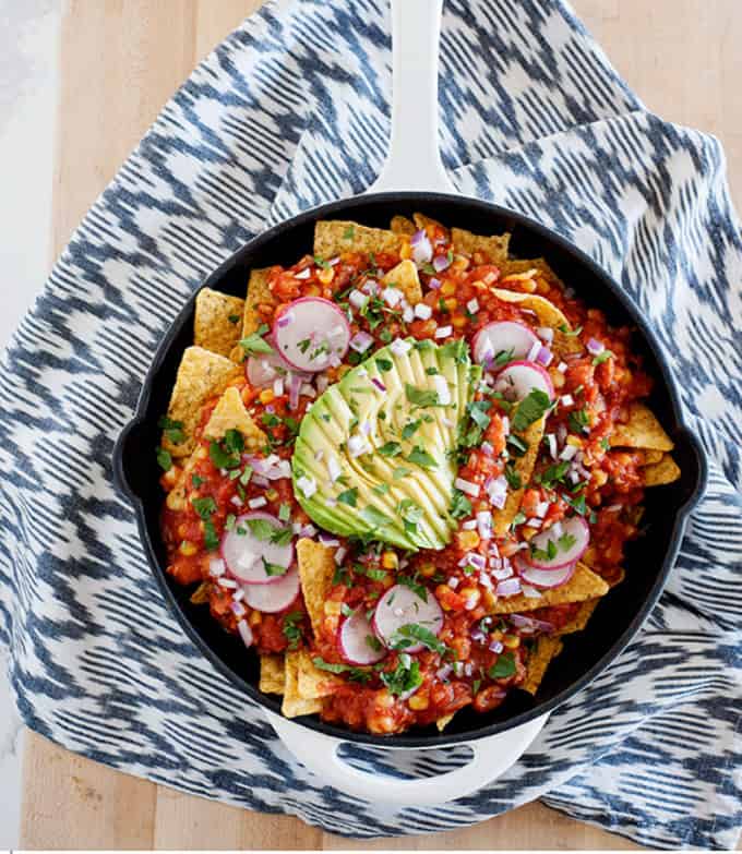 Skillet with tortilla chips topped with tomato sauce, radishes, chickpeas, and avocado