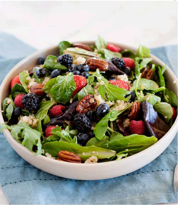 Green salad, in a white bowl, topped with mixed berries and toasted pecans.