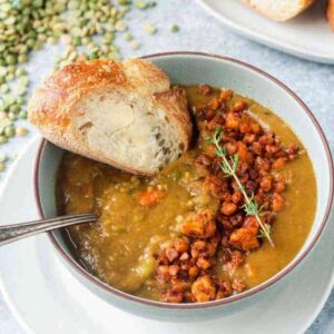 Bowl of vegan split pea soup topped with tempeh crumbles and served with a side of buttered bread.