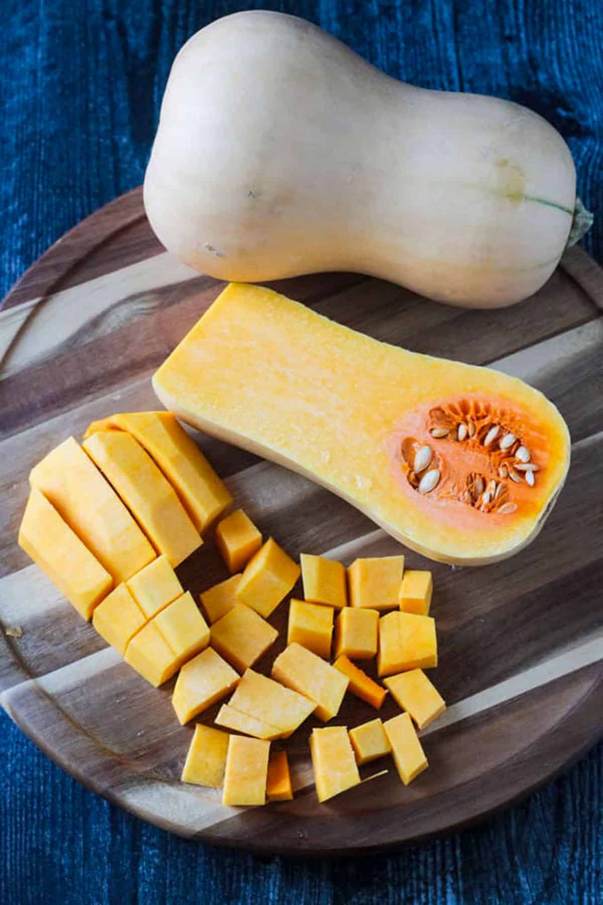 One whole butternut squash, one half butternut squash cut side up, and cubed pieces of butternut squash on a round wooden cutting board.