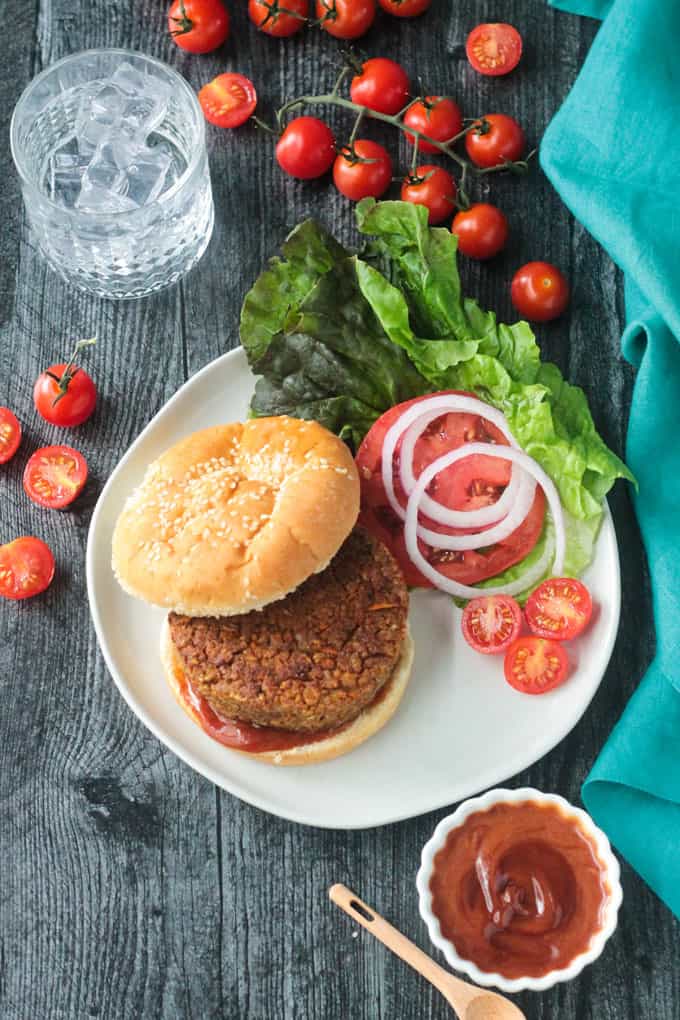 Plate with a burger patty on a bun next to garnishes of lettuce, tomoto, and red onion.