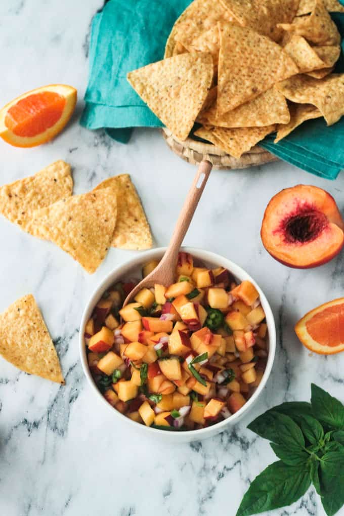 Bowl of peach salsa on a marble slab with tortilla chips scattered around it.