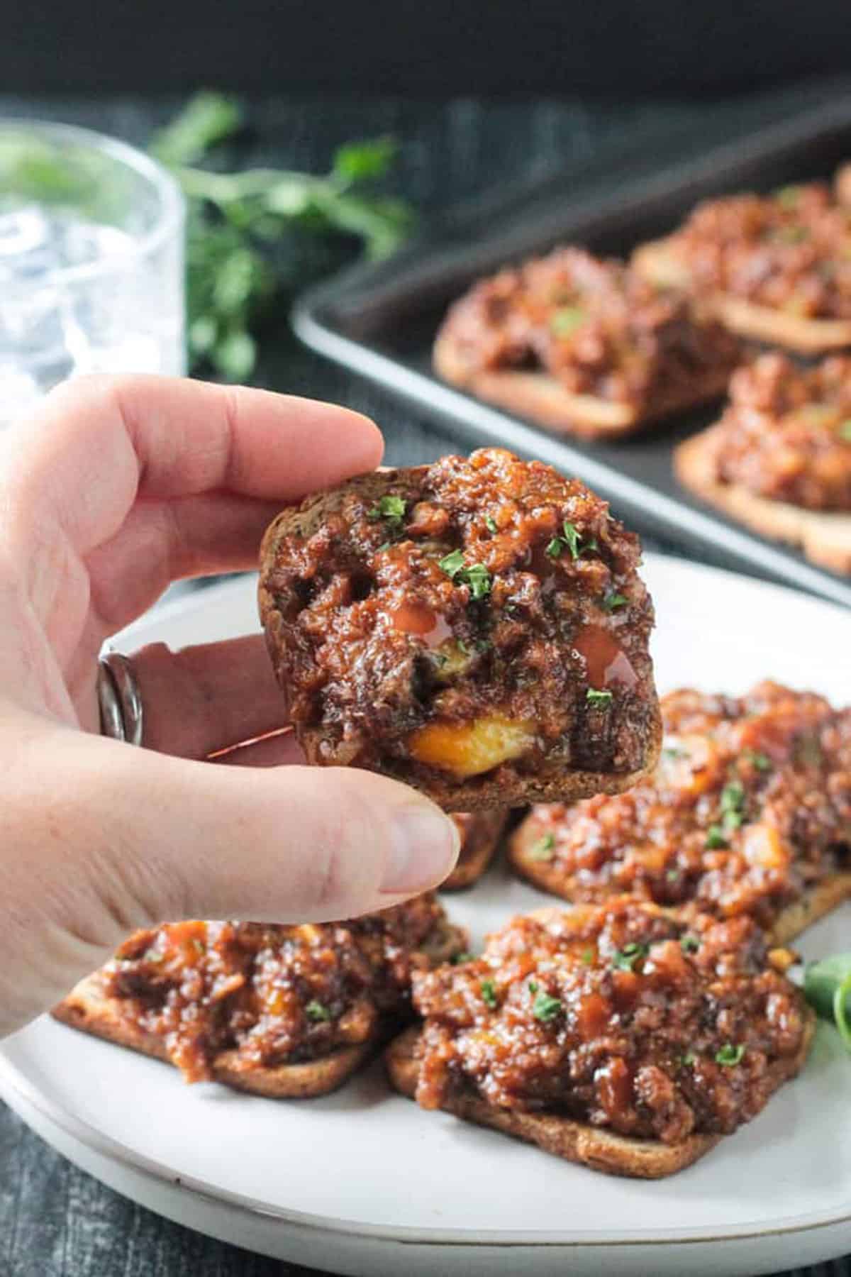 Hand holding one small square rye toast baked with meat and cheese.