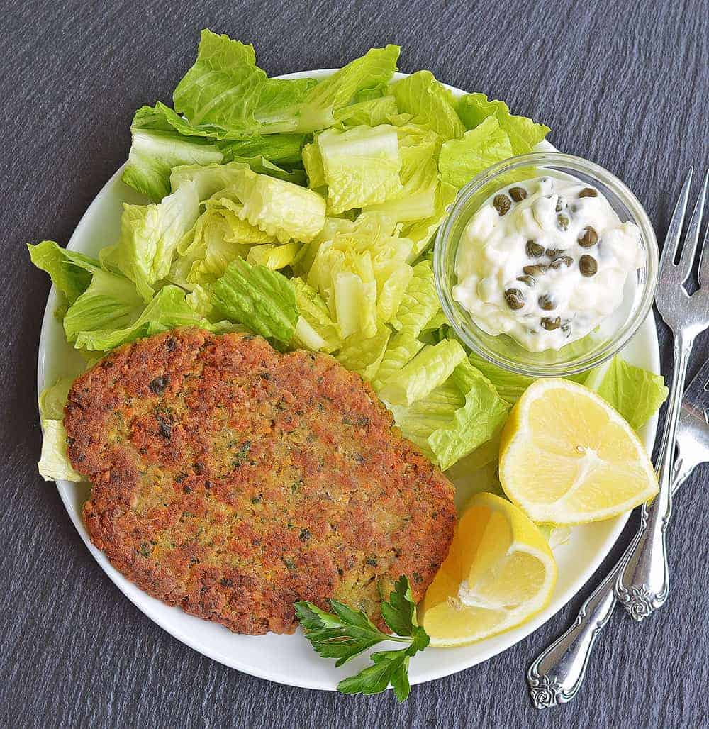 chickpea cutlet on a plate with lettuce, a tiny jar of sauce, and two lemon wedges