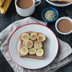 Breakfast toast on a white plate surrounded by a cup of coffee, bunch of bananas, small bowl of almond butter, and more toast slices.