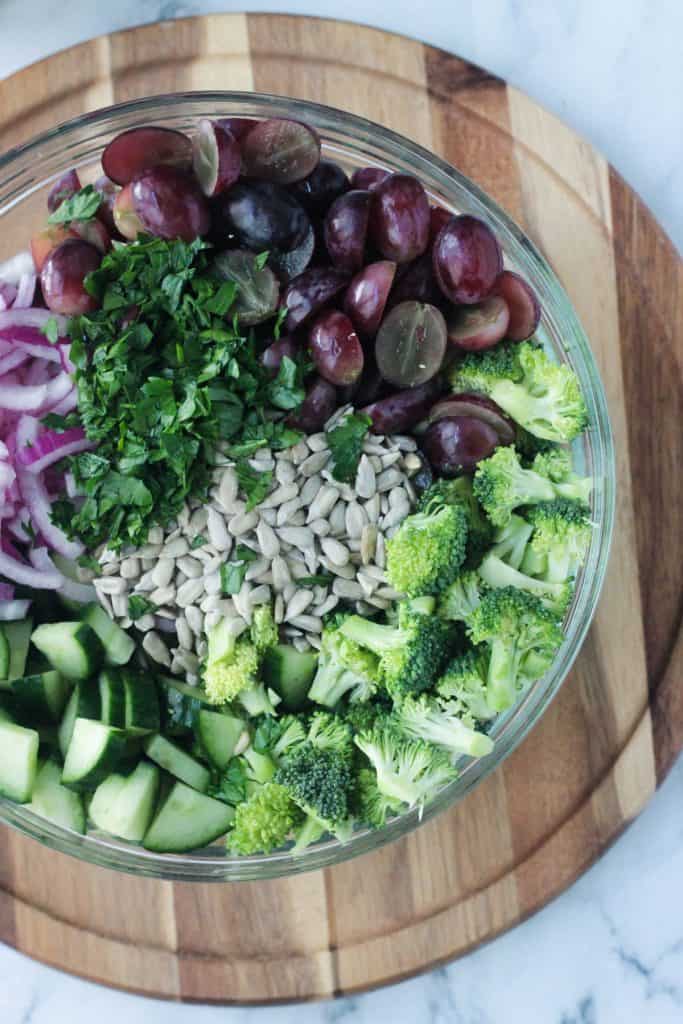 Close up of broccoli, grapes, cucumber, red onion, parsley, and sunflower seeds in a glass bowl.