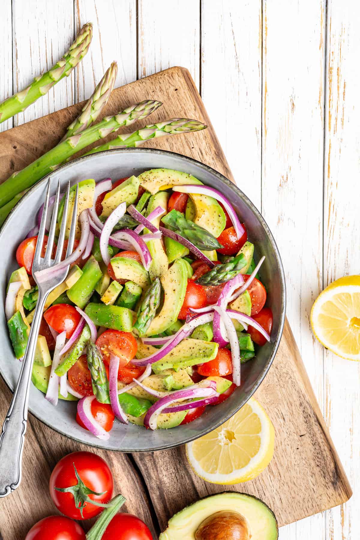Asparagus, avocado, tomato, and red onion salad in a bowl on a wooden board.