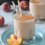 Two peach slices in front of smoothie in a glass with two straws.