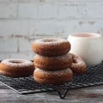 Stack of three cider donuts on a black wire cooling rack in front of a coffee mug.