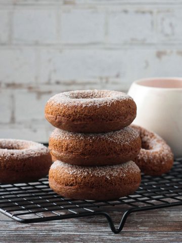Stack of three cider donuts on a black wire cooling rack in front of a coffee mug.