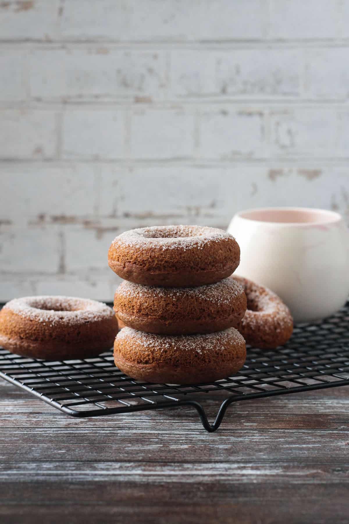 Stack of three cider donuts on a black cooling rack in front of a mug.