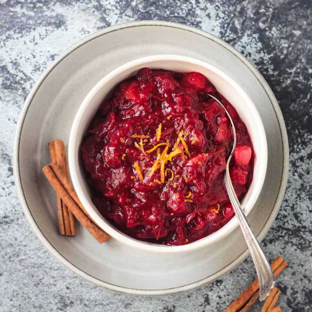 Cranberry sauce in a white bowl on a white plate with 2 cinnamon sticks.