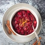 Cranberry sauce in a white bowl on a white plate with 2 cinnamon sticks.