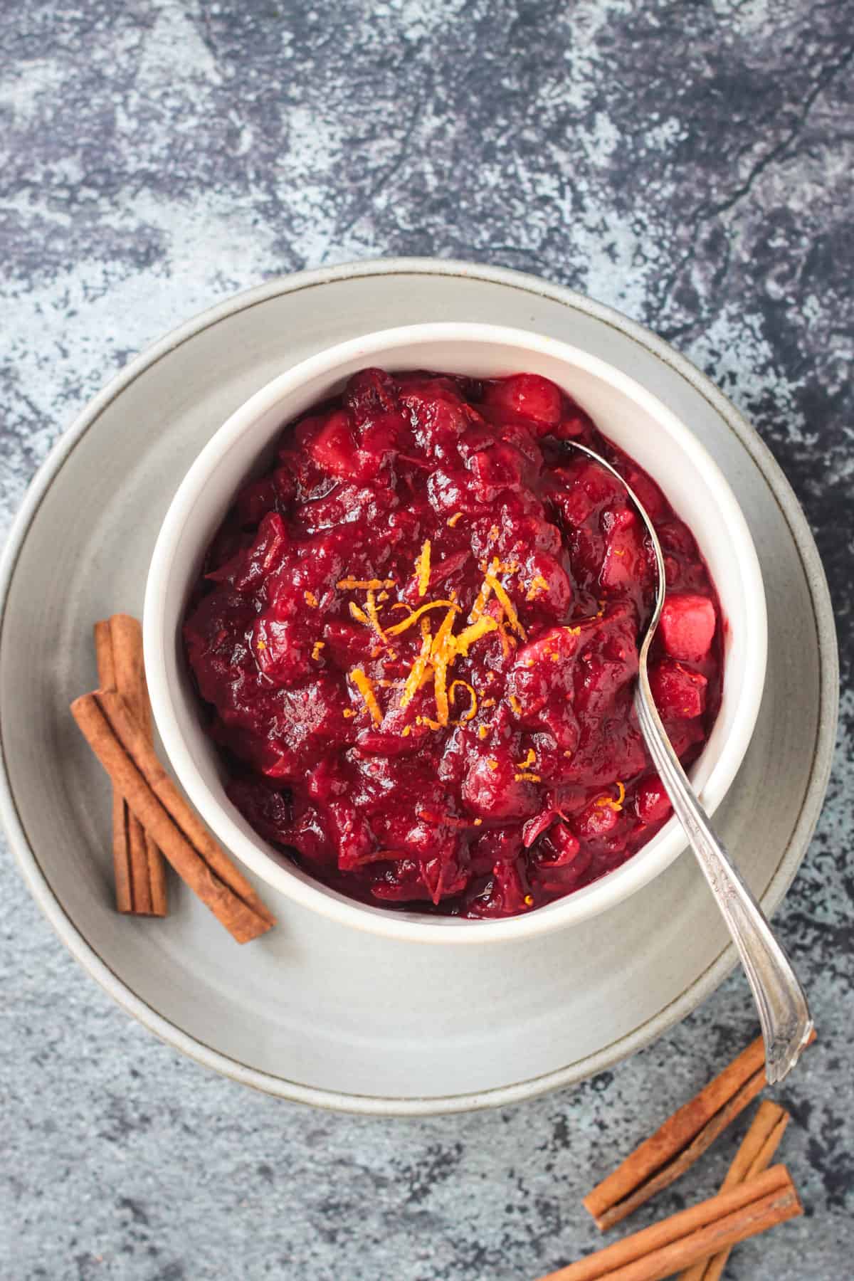 Spiced orange cranberry sauce in a white bowl on a white plate with 2 cinnamon sticks.