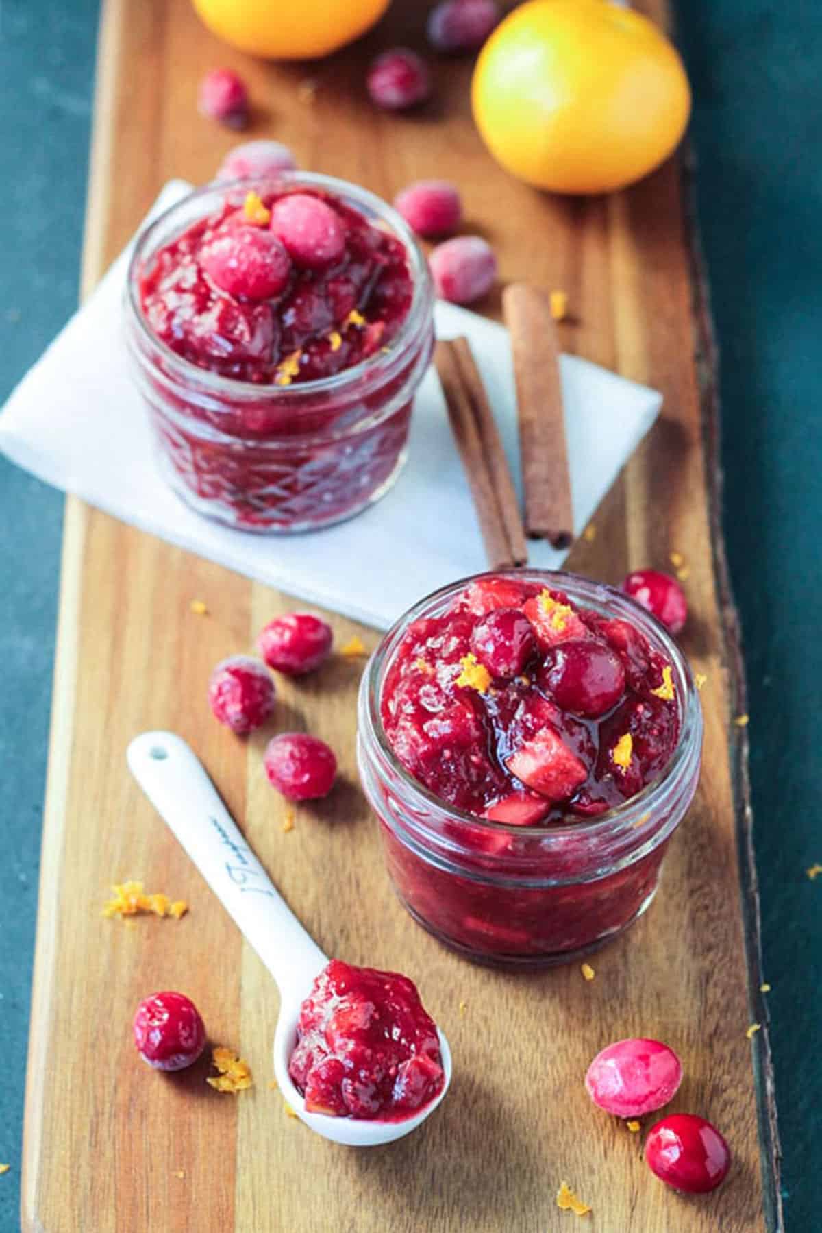 Two small jars of sauce on a wooden serving board with fresh cranberries scattered about.