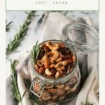 Open jar of spiced roasted nuts surrounded by fresh rosemary sprigs.