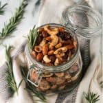 Open jar of spiced roasted nuts surrounded by fresh rosemary sprigs.