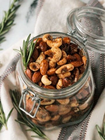 Open jar of spiced roasted nuts surrounded by fresh rosemary sprigs.