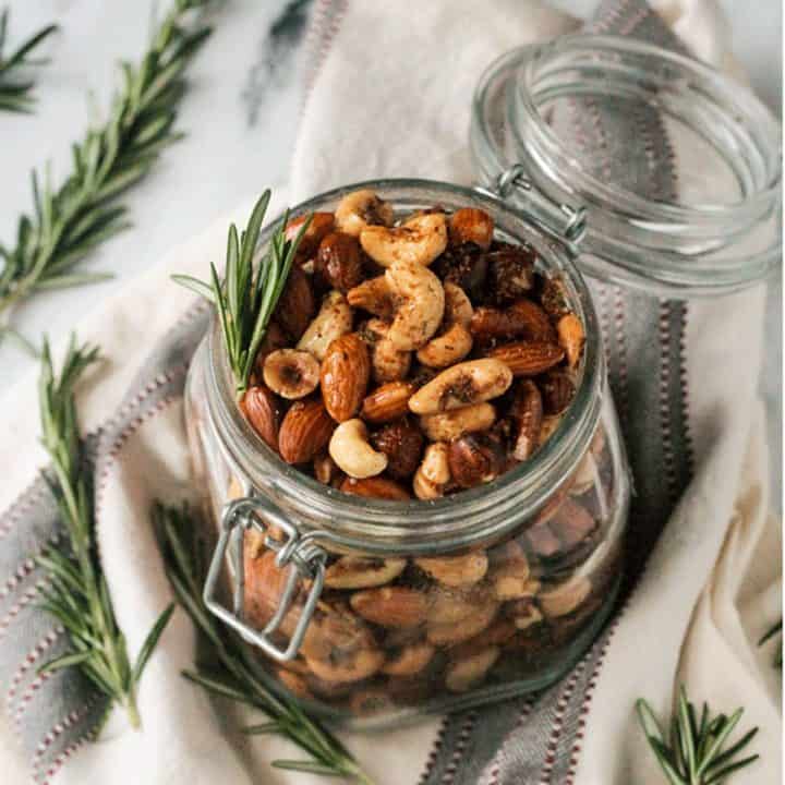 Open jar of spiced roasted nuts surrounded by fresh rosemary sprigs.