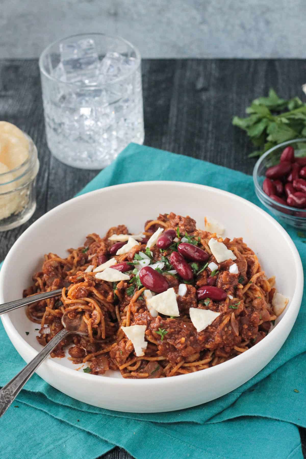 Crushed crackers and kidney beans on top of chili spaghetti in a white flat bowl.