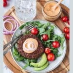 Burger patty topped with sauce and raw onion next to arugula and sliced avocado on a plate.