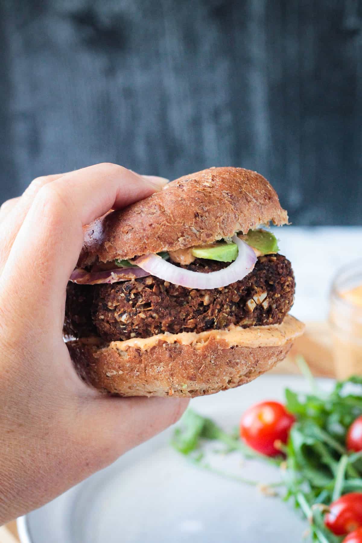 Hand holding a burger on a whole wheat bun with sauce, avocado and onion slices.