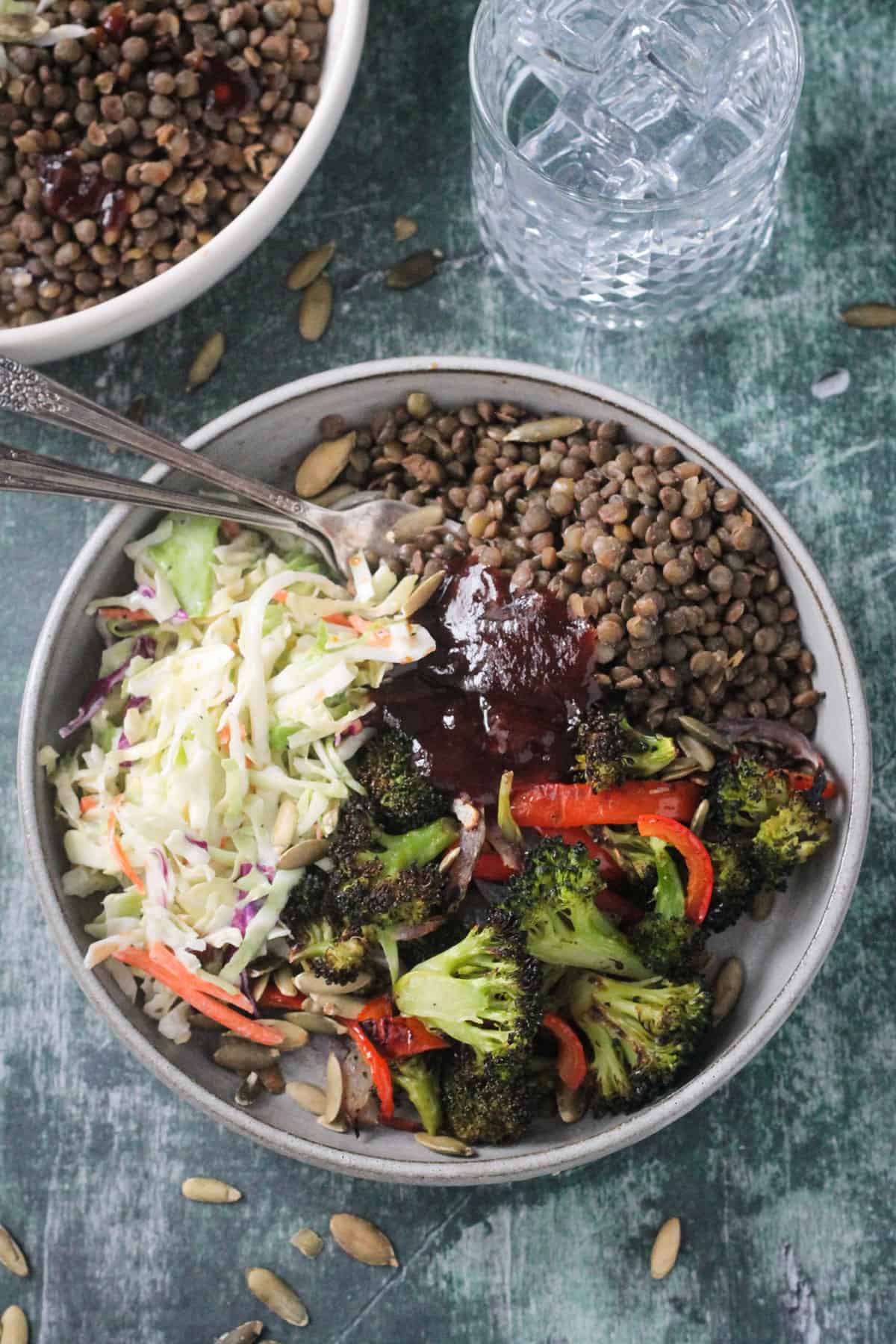 Two forks in a pile of lentils next to roasted broccoli, coleslaw, and bbq sauce.
