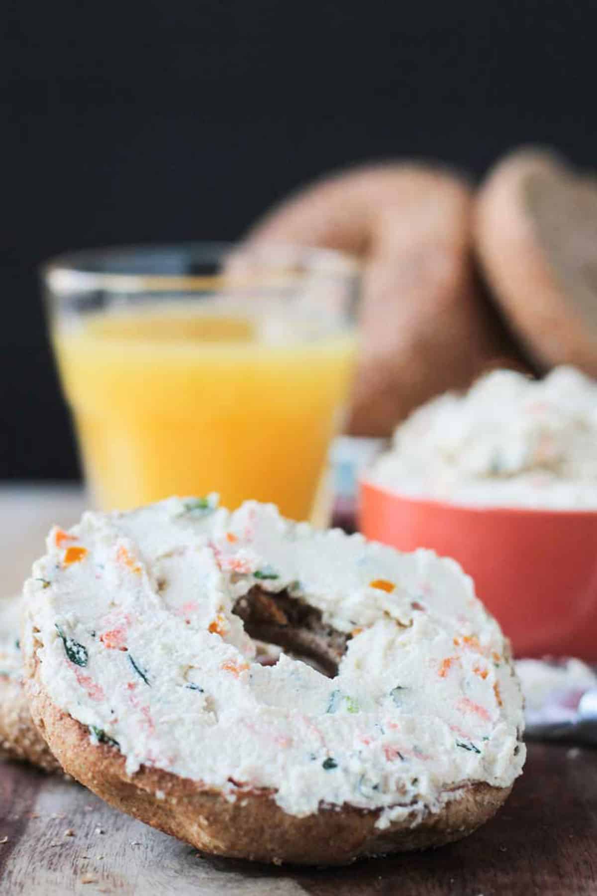 Veggie cream cheese spread on a bagel half in front of a glass of orange juice.